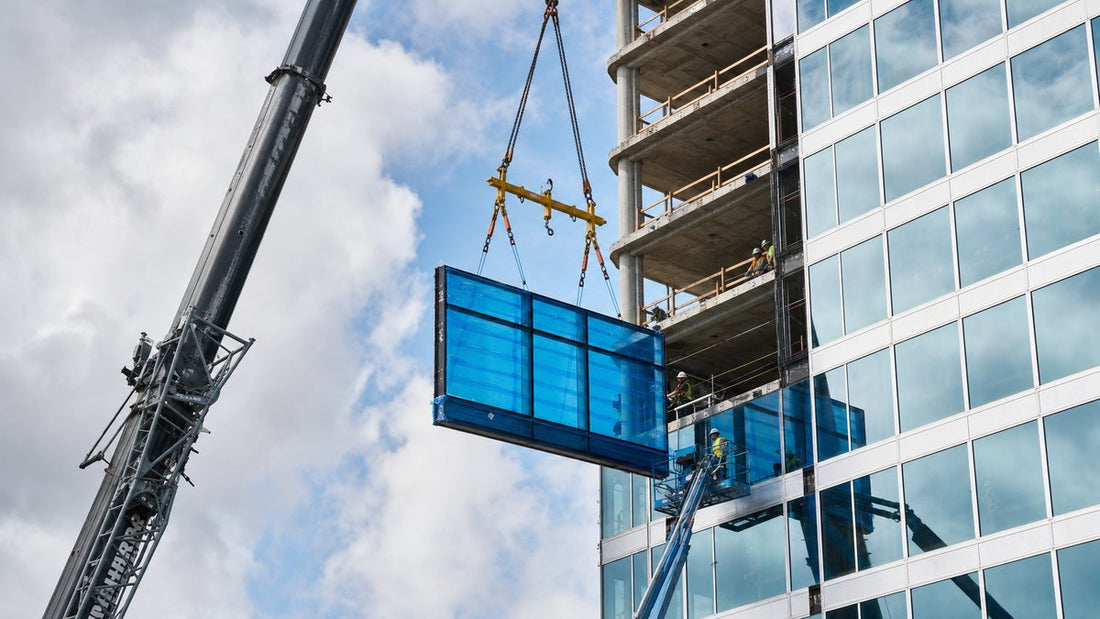 Crane hoisting a large architectural glass cladding panel wrapped in blue protective film at a high-rise construction site