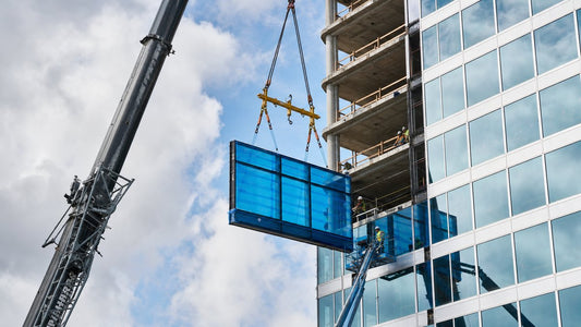 Crane hoisting a large architectural glass cladding panel wrapped in blue protective film at a high-rise construction site
